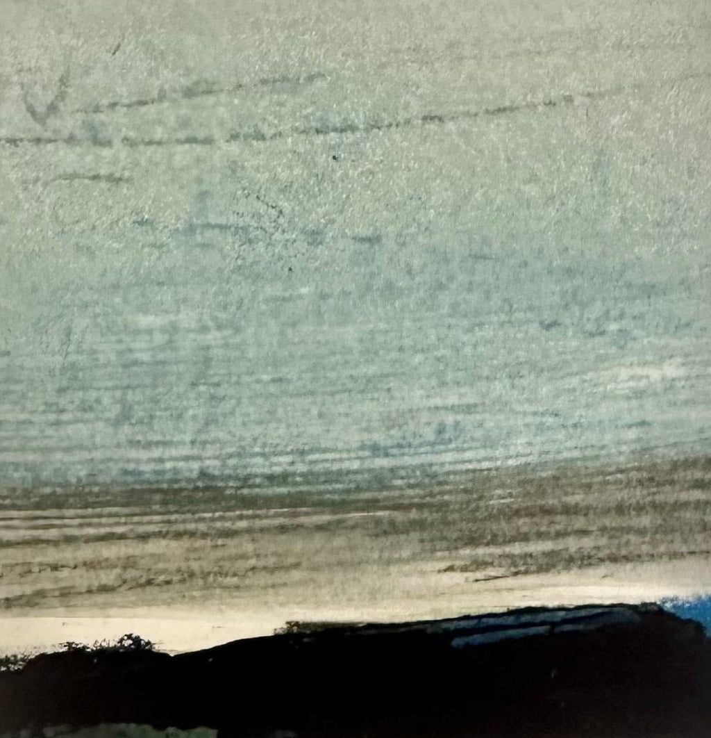 A hand holding a square white matted art print featuring a small abstract landscape painting with a light blue sky, gray middle band, and dark black landscape at the bottom.