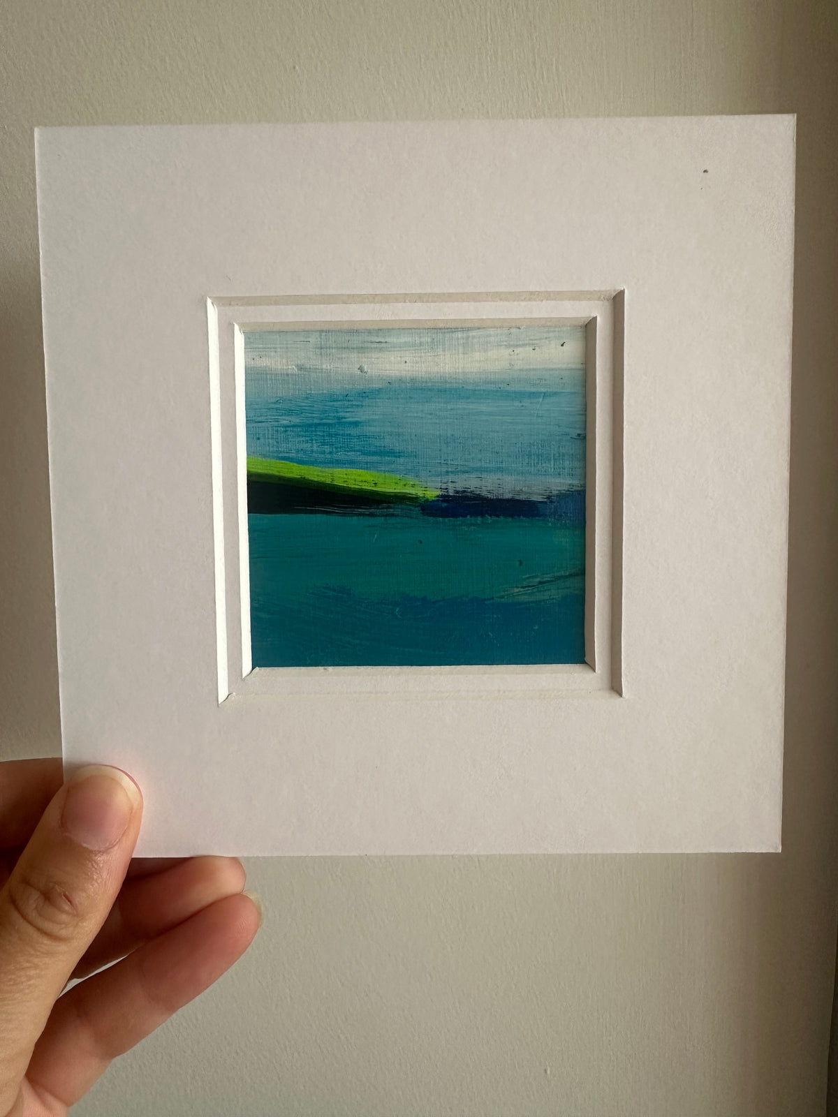 A hand holding a square white matted art print featuring a small abstract landscape painting with blue, teal, and lime green brushstrokes, set against a neutral beige wall.