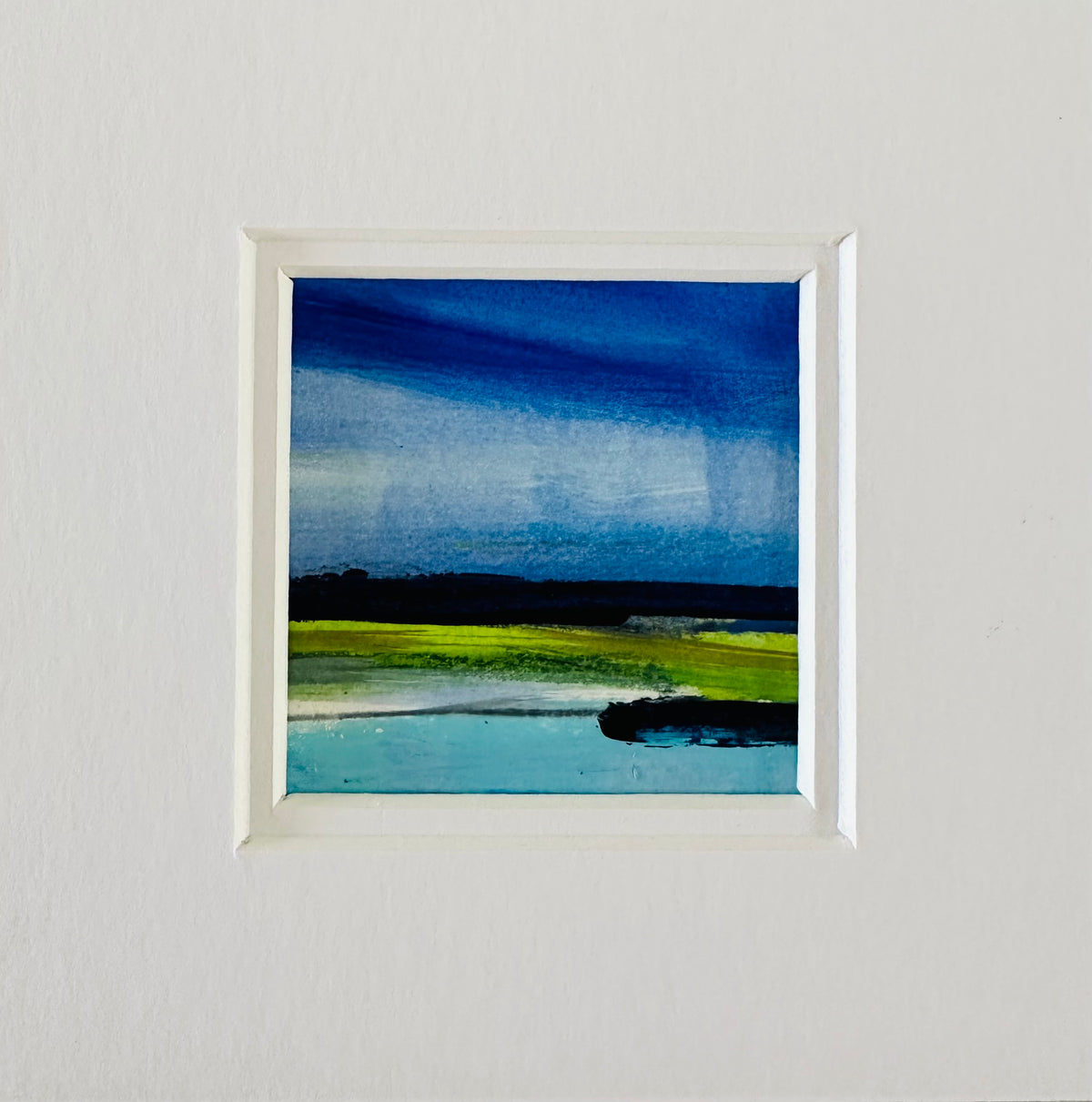 A square framed abstract landscape painting featuring deep blue water, a bright green shoreline, and a dark landmass under a stormy blue sky, mounted on a textured white wall.