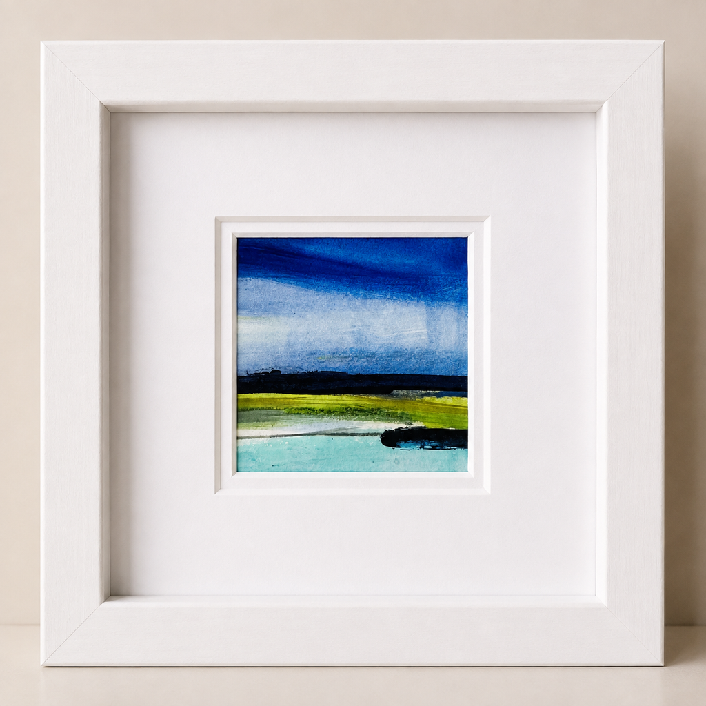 A square white framed art print featuring a landscape painting with a deep blue sky, green fields, and turquoise water, set against a neutral beige background.