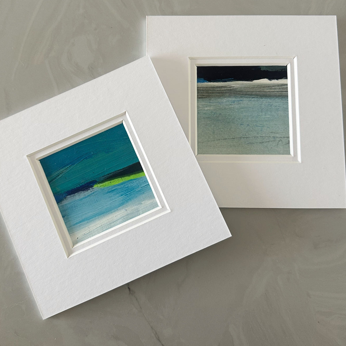 Two matted art prints with white mats are displayed on a gray marble surface. One print features an abstract painting in shades of blue, teal, and green, while the other shows a seascape painting with blue water and a dark sky.